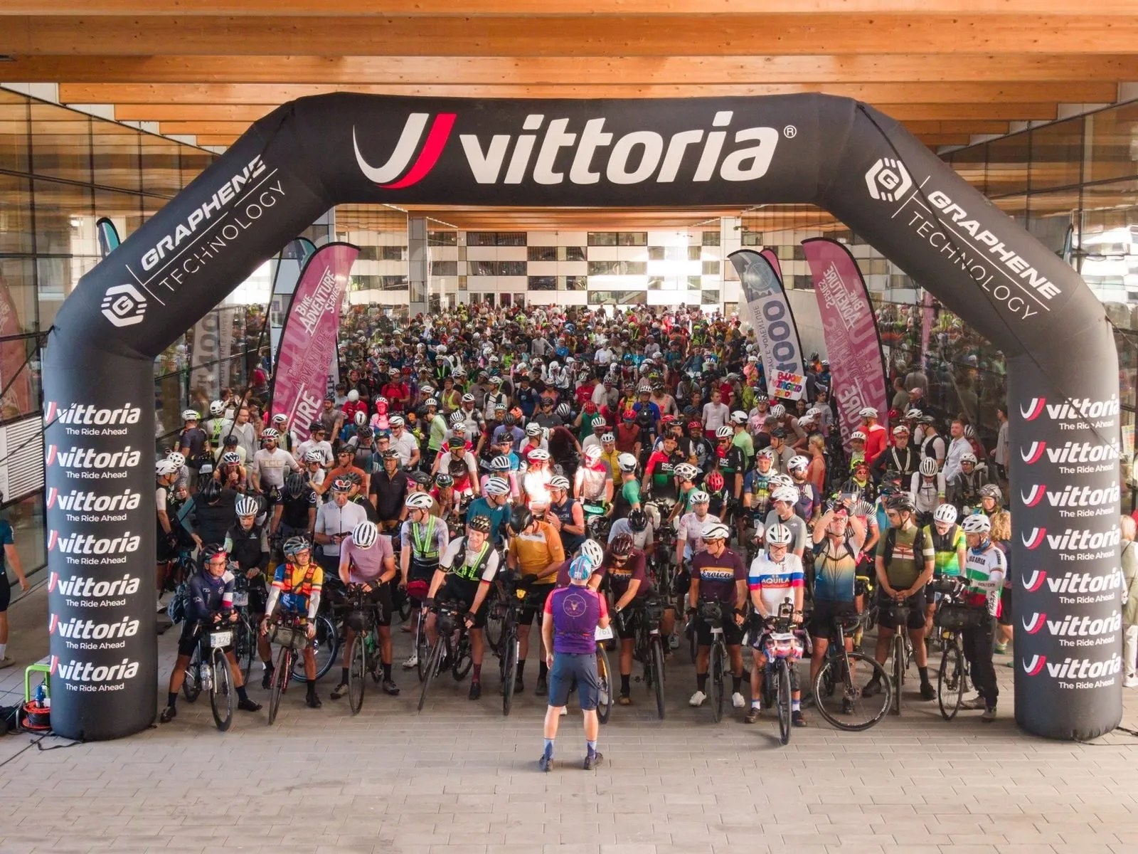 Centinaia di ciclisti con le bici cariche sotto l’arco di partenza di uno sponsor